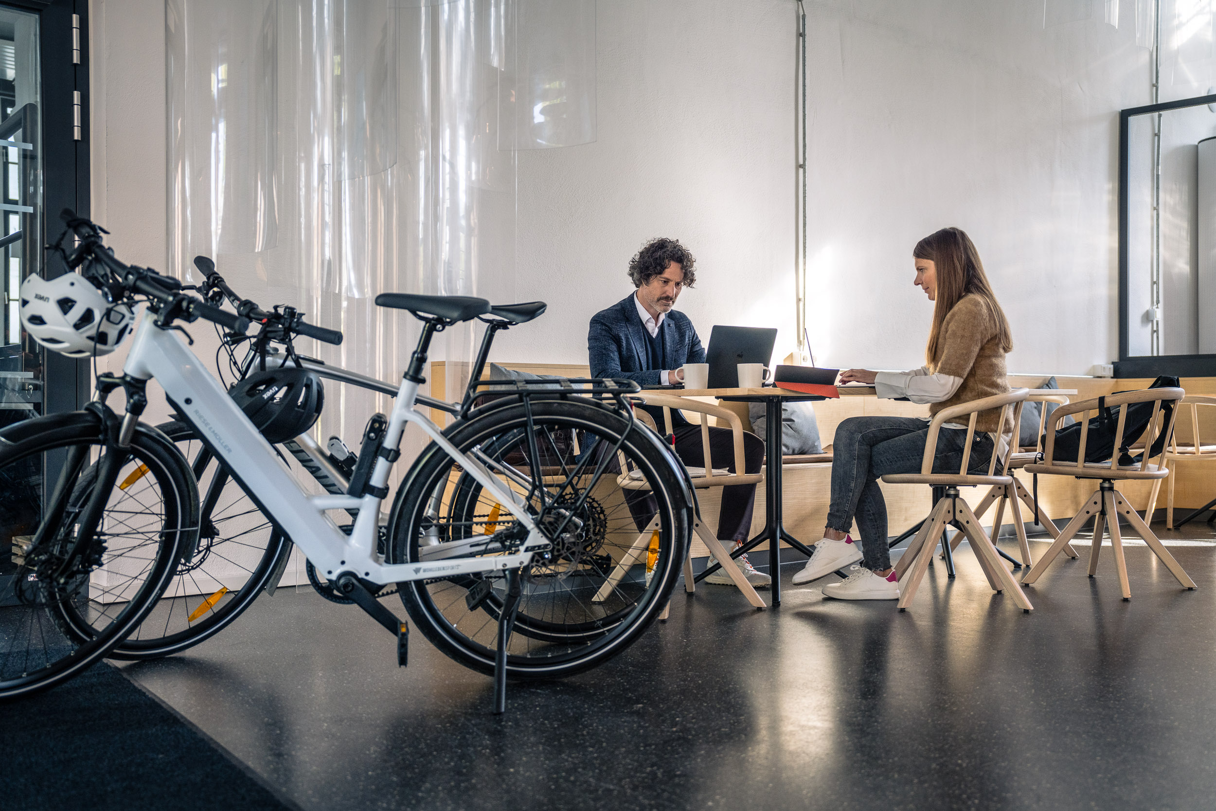 Zwei Personen arbeiten an einem Tisch mit Laptops in einem hellen Raum, während im Vordergrund mehrere geparkte Fahrräder stehen.