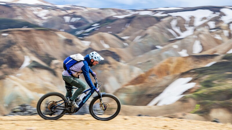 Ein Mountainbiker mit Helm fährt zügig über einen Schotterkamm, im Hintergrund bunte Berge mit Schneefeldern.